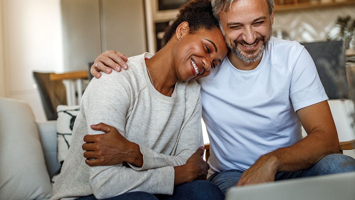 A smiling couple sitting on a couch while viewing a laptop in their home