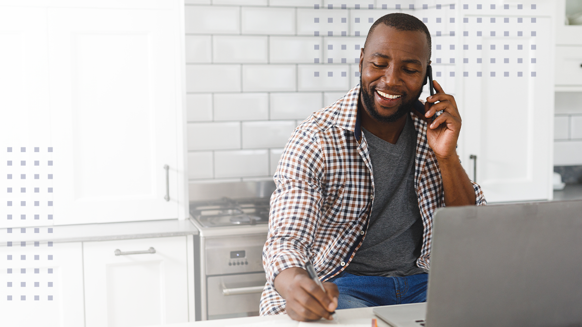 A man talking on a cell phone in front of a laptop while taking notes in a kitchen