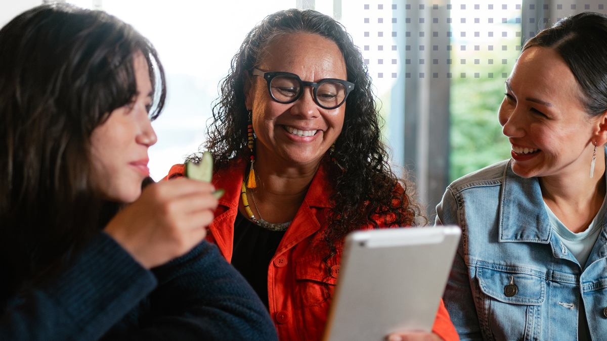 A group of 3 women smiling together while looking at a tablet