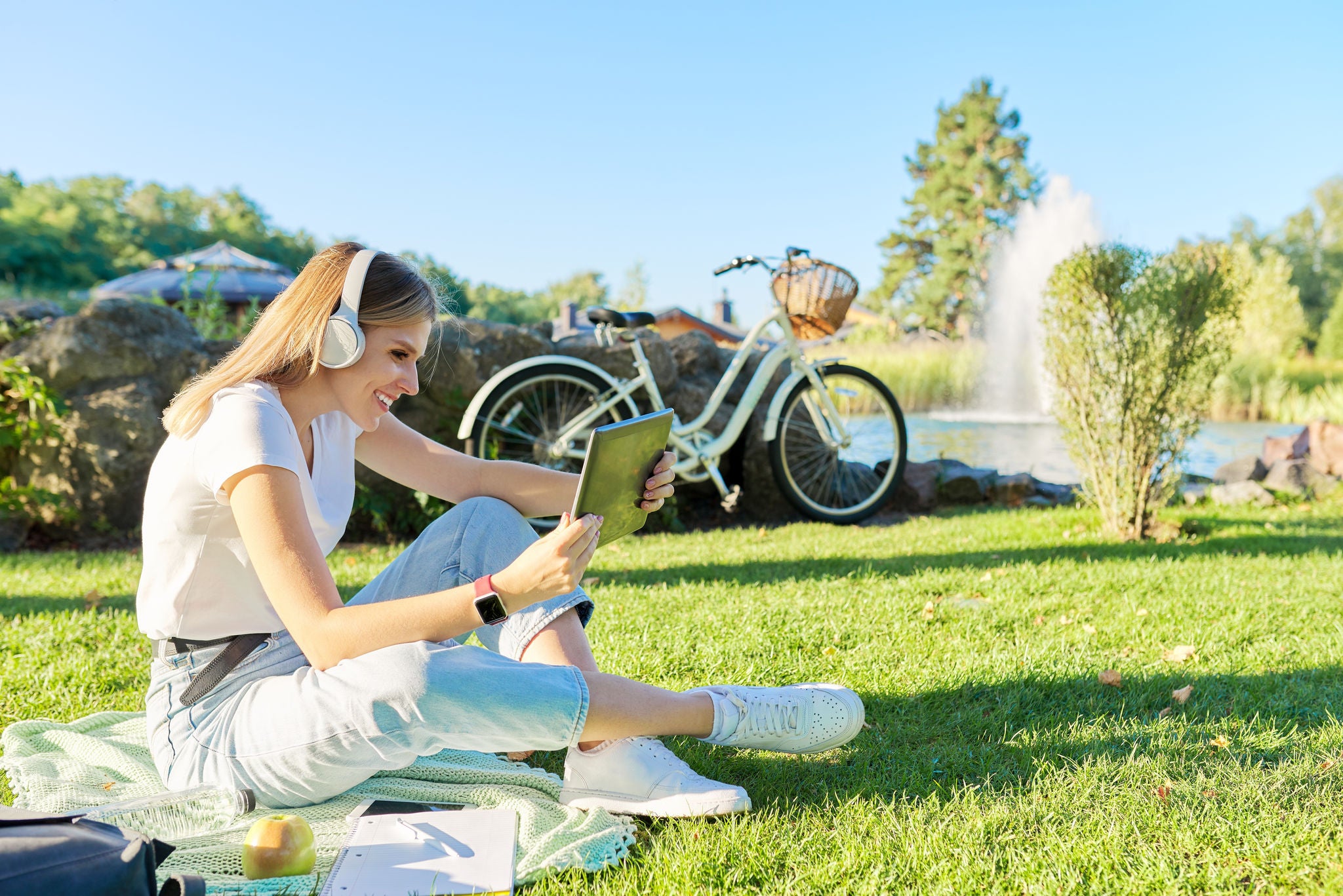 Young woman in headphones with digital tablet looking in screen of gadget, video, film, call. Female sitting on green lawn in park on sunny summer day