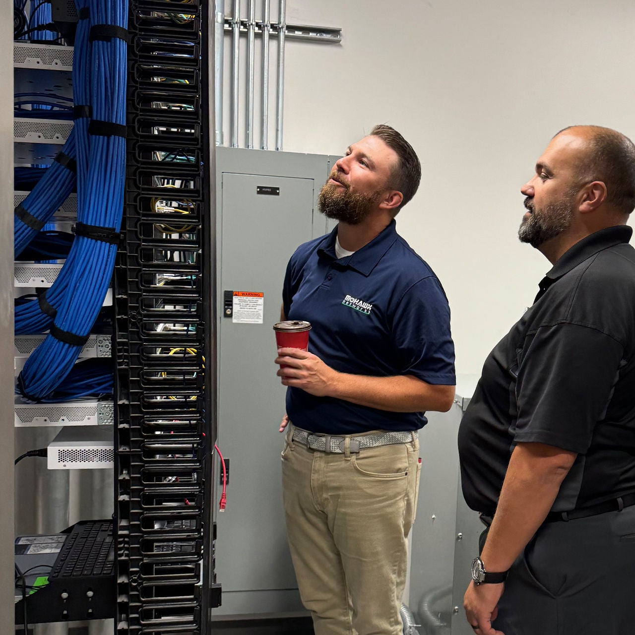 Two men observing a network server rack with wiring