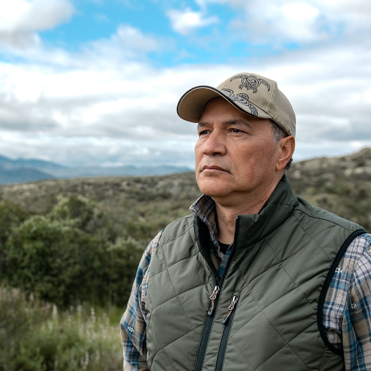 A tribal man with a hat looking off into the distance outdoors by a mountain range