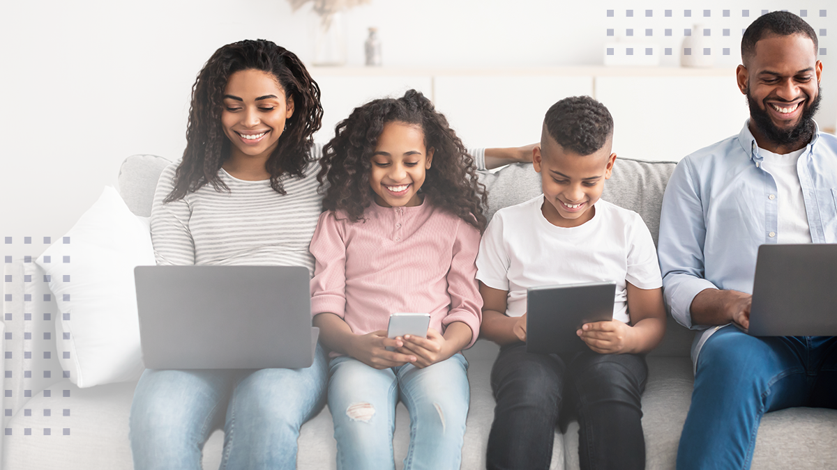 A smiling family all sitting on a couch while using their electronic devices